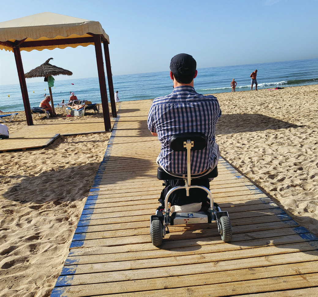 Mann im Rollstuhl steht auf Holzbrücke und schaut ins Meer 